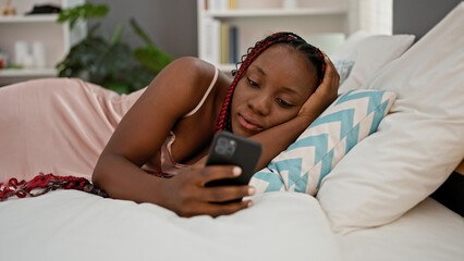 Beautiful african american woman at home, comfortably relaxing on bed in pajamas, typing message...