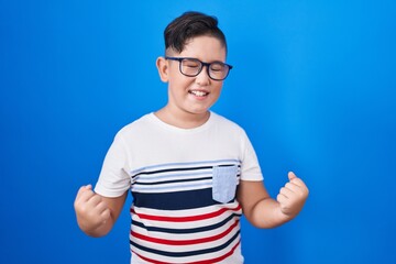 Young hispanic kid standing over blue background very happy and excited doing winner gesture with...