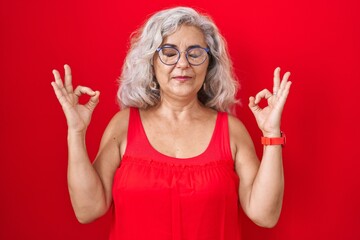 Middle age woman with grey hair standing over red background relaxed and smiling with eyes closed doing meditation gesture with fingers. yoga concept.