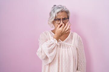 Middle age woman with grey hair standing over pink background smelling something stinky and disgusting, intolerable smell, holding breath with fingers on nose. bad smell - obrazy, fototapety, plakaty