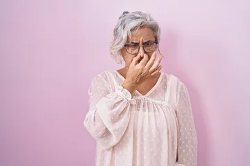Foto op Canvas Middle age woman with grey hair standing over pink background smelling something stinky and disgusting, intolerable smell, holding breath with fingers on nose. bad smell © Krakenimages.com