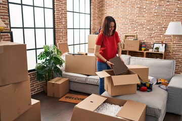 Young beautiful hispanic woman smiling confident unpacking cardboard box at new home