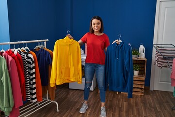 Young beautiful hispanic woman smiling confident holding clothes on rack at laundry room