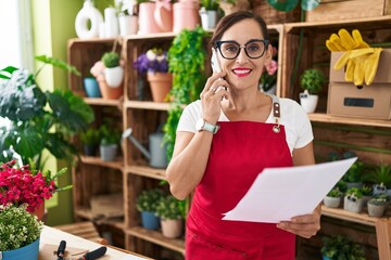 Young beautiful hispanic woman florist talking on smartphone reading document at flower shop