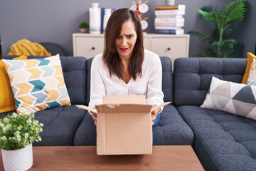 Middle age brunette woman opening cardboard box clueless and confused expression. doubt concept.