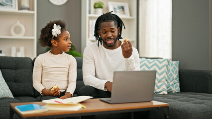African american father and daughter sitting on sofa eating croissant drawing and working at home