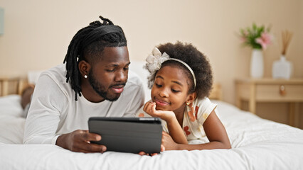 African american father and daughter watching video on touchpad lying on bed at bedroom - Powered by Adobe