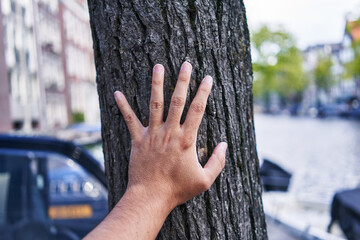 A man's hand gently touches a tree trunk on a vibrant city street, expressing connection with...