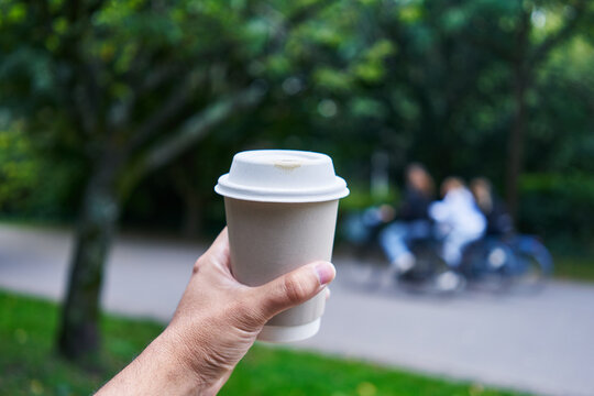 A man's hand holding a coffee cup with people blurred in the background outdoors.