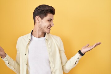 Young hispanic man standing over yellow background smiling showing both hands open palms,...