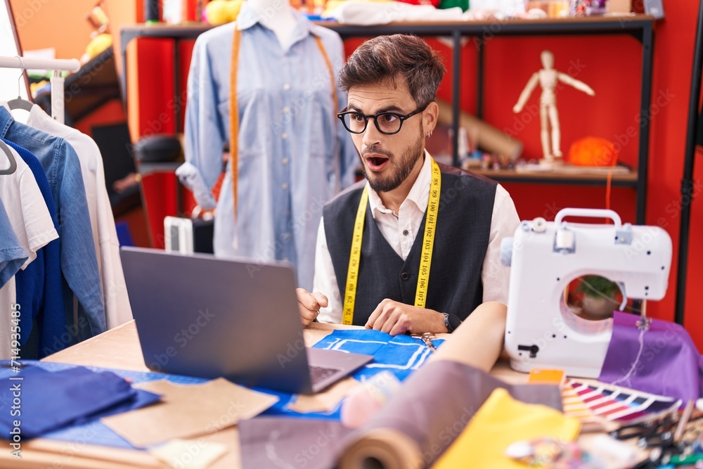 Sticker young hispanic man with tattoos dressmaker designer using laptop at tailor room scared and amazed wi