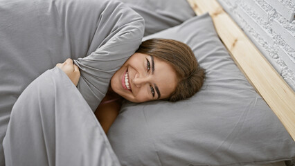 A confident, smiling young hispanic woman enjoys a cozy, comforting morning resting in bed, basking...
