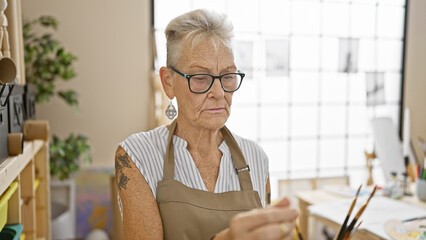 Dedicated senior grey-haired woman artist, holding paintbrushes, deeply engrossed in painting at...