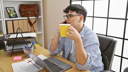 Young man talking on phone while holding coffee mug at his cluttered office desk with laptop and...