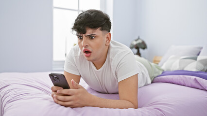 A shocked young man lies on a bed in a modern bedroom, holding a smartphone with an astonished expression.