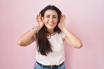 Fototapeta na wymiar Young brunette woman standing over pink background trying to hear both hands on ear gesture, curious for gossip. hearing problem, deaf