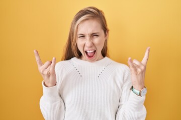 Young caucasian woman wearing white sweater over yellow background shouting with crazy expression doing rock symbol with hands up. music star. heavy music concept. - Powered by Adobe