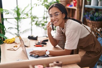 Young beautiful hispanic woman florist talking on smartphone using laptop at florist