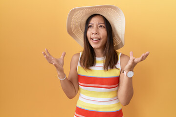 Middle age chinese woman wearing summer hat over yellow background crazy and mad shouting and yelling with aggressive expression and arms raised. frustration concept.