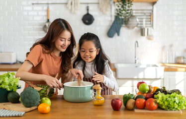 Portrait of happy love asian family mother with little asian girl daughter child help cooking food healthy eat with fresh vegetable testing smell soup in a pot with spoon.help mom in kitchen at home - obrazy, fototapety, plakaty
