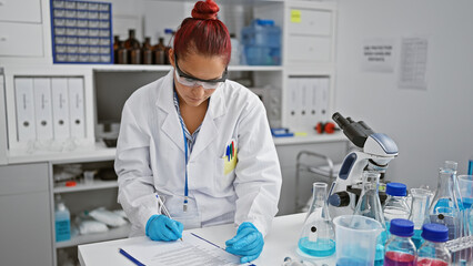 Focused young, redhead woman scientist engrossed in medical research, taking serious notes at lab...