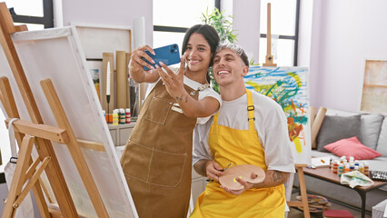 A woman and man artist couple take a selfie in a vibrant painting studio, surrounded by canvases...