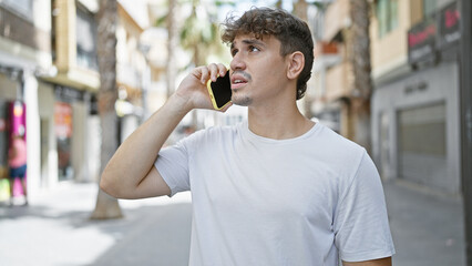 Cool-looking, handsome young hispanic man seriously engaged in conversation on his smartphone, standing under the sunny urban street's casual fashion backdrop with a concentrated expression.