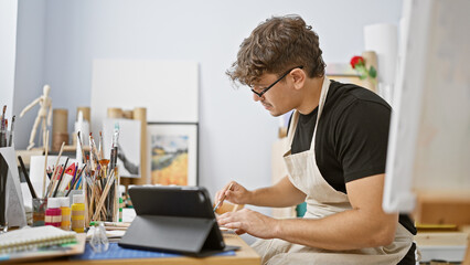 Attractive young hispanic man, a committed art student, fervently engrossed in drawing on his touchpad and notebook at his studio - obrazy, fototapety, plakaty