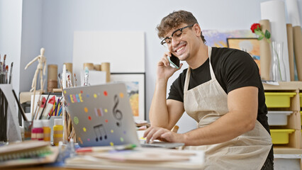 Handsome young hispanic artist, deep in conversation on smartphone, skillfully using laptop at his...