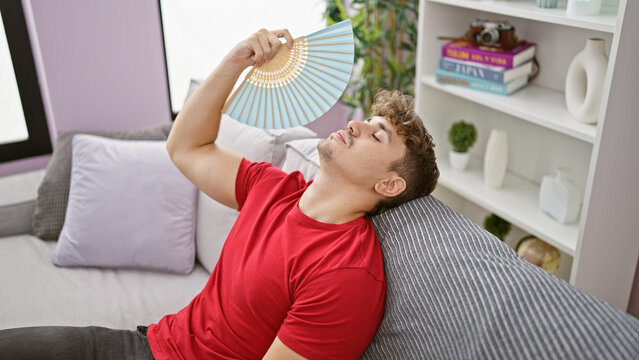 Hot weather woes, young hispanic man suffers, fanning himself with handfan sitting on sofa at home, exposing the struggles of indoor heat