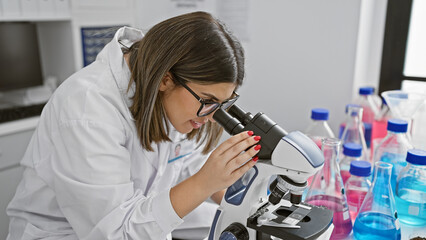 A focused hispanic woman scientist examines samples under a microscope in a well-equipped laboratory.
