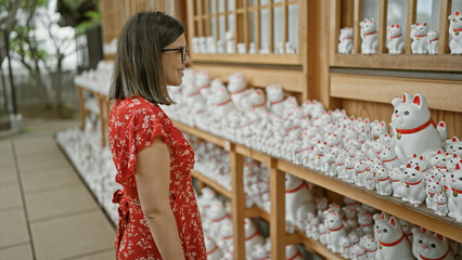 Beautiful hispanic woman in glasses counting on luck, standing by the famous lucky cat statue at gotokuji temple in tokyo