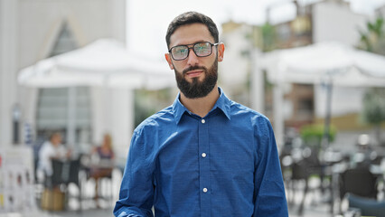 Young hispanic man business worker standing with serious face at coffee shop terrace