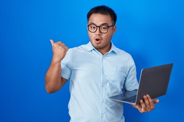 Chinese young man using computer laptop surprised pointing with hand finger to the side, open mouth amazed expression.