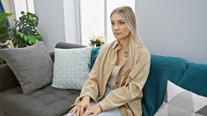 Beautiful, worried blonde woman, a young adult, sitting on the indoor sofa at home - pondering,...