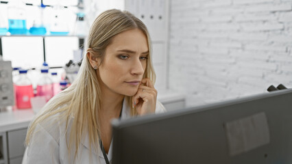 Serious, beautiful young blonde woman scientist thinking hard while working on computer, conducting research in medical lab