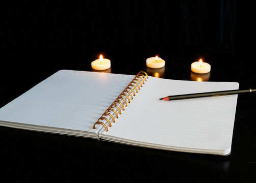 Cahier à spirale ouvert sur une page blanche avec un crayon sur une table noire avec trois petites bougies allumées 