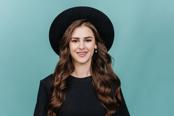Smiling woman in a black outfit and hat, isolated on a teal background