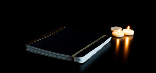 Cahier fermé sur une table noire avec 3 petites bougies chauffe plat allumées et leur reflet -...
