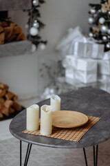 Three white candles of varying heights on a round wooden plate, placed on a dark table. A decorated Christmas tree with white ornaments, lights, and wrapped presents forms the festive background.