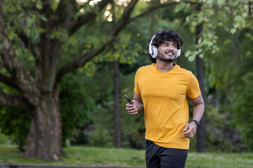Fototapeta premium Young smiling Indian man running in the park wearing headphones and holding a mobile phone