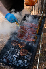 Basting succulent mackerel fillets on a grill over charcoals, with burger patties and rising smoke in a serene forest