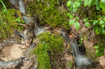 Stream of flowing water with motion blur, finds its way between the moss and grass-covered rocks. Background image