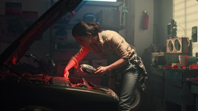 Beautiful woman looks under hood of car indoors. Young female automobile mechanic repairing car engine in automotive workshop with wrench and lamp. Car service and maintenance. Repair service.