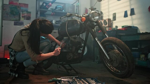 Beautiful young woman in casual clothes using spanner while repairing motorcycle in repair shop. Female bike repair station worker fixing motorcycle in garage. Mechanical hobby and repairs concepts.