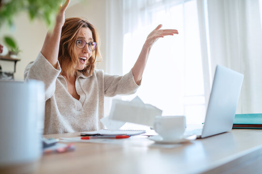 Beautiful middle-aged woman in glasses extremely surprised with new higher taxes watching, throwing bills at laptop in home office. Small business troubles, bull market or money savings concept image