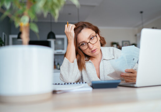 Beautiful middle-aged pensive woman in glasses with monthly bills thinking about family home budget. Small business, home finances, money savings concept image.