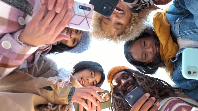Low angle view of multiethnic group of friends in circle using their smart mobile phones together outdoors.
