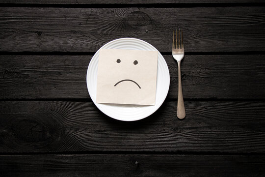 A white plate and paper with a drawn breast stand on a black wooden table and a fork next to it, nothing to eat, hunger