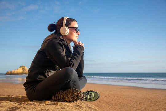 Woman Sitting On A Rock Looking Out To Sea, Listening To Music On Headphones, Winter Sun.